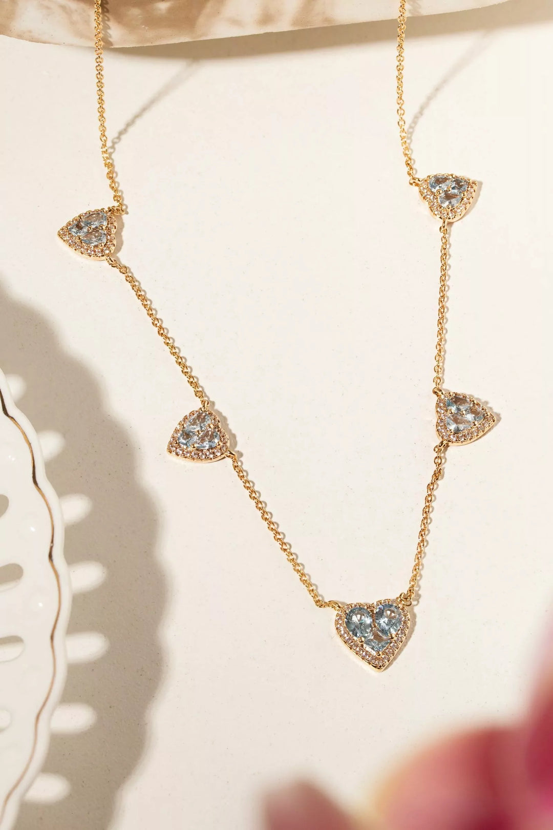A delicate gold chain necklace featuring five small heart-shaped puffed charms, each adorned with clear pavé stones and a central light blue heart, displayed on a soft cream background.