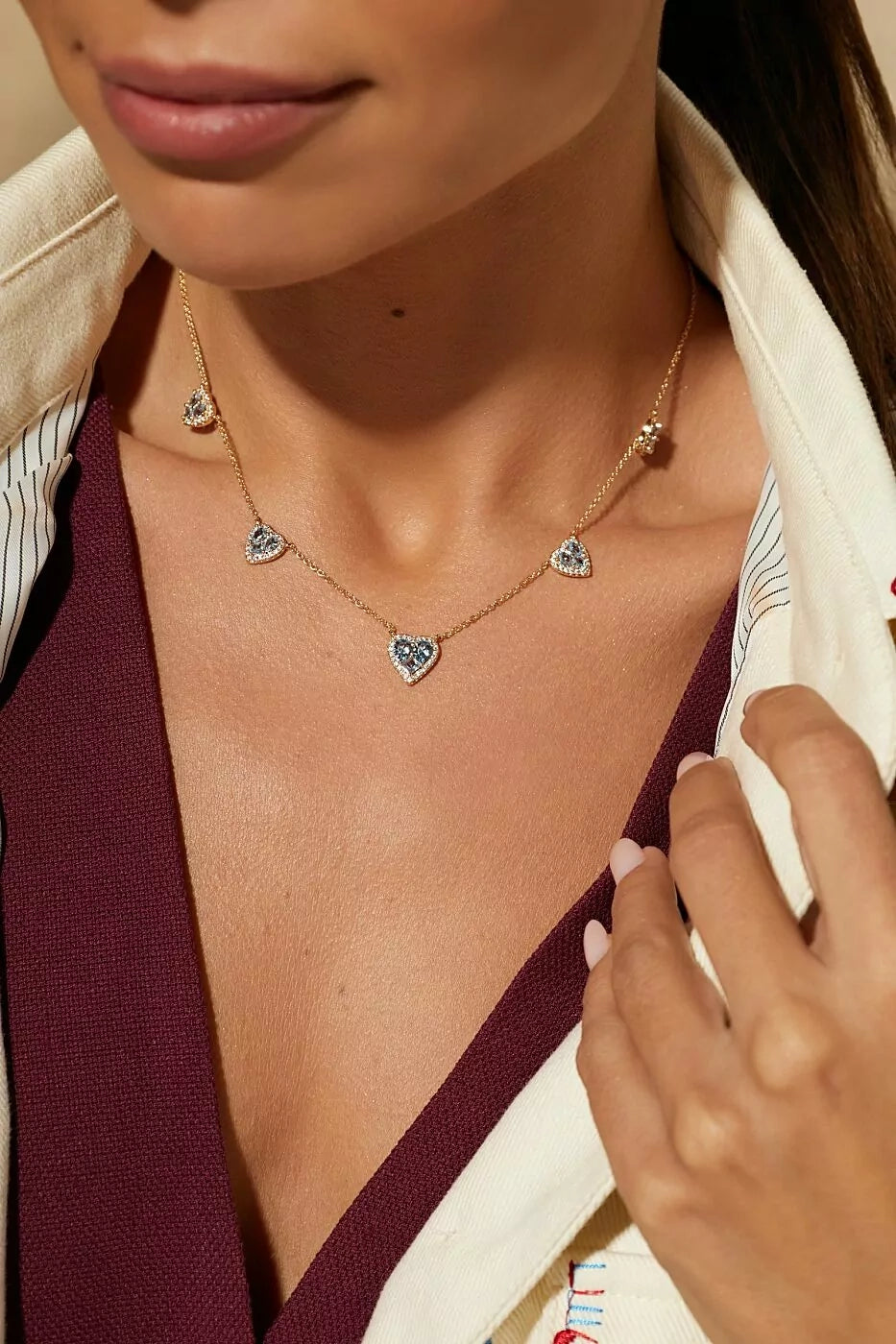 A close-up lifestyle shot of a model wearing a delicate gold chain necklace with three small heart-shaped charms, each adorned with sparkling clear stones. The necklace is styled with a cream-colored collared shirt and a deep burgundy top.