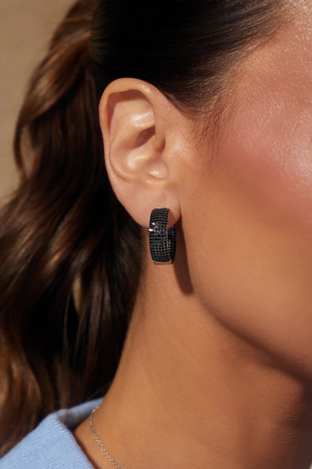 A close-up side profile of a woman wearing a small black textured hoop earring, with her ear, hair, and neck visible against a warm neutral background.