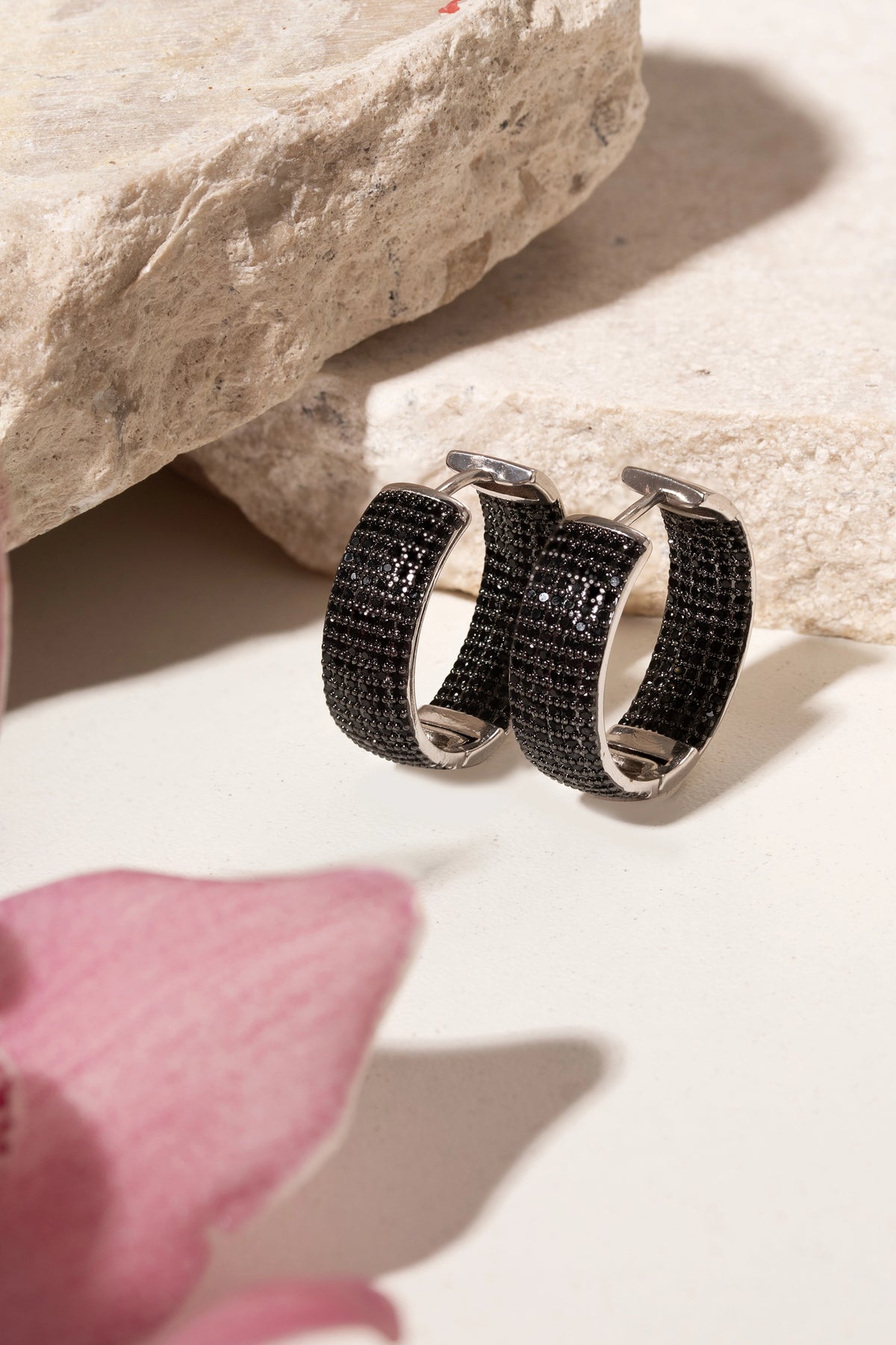 A pair of silver hoop earrings with textured black woven detailing, displayed on a light beige surface next to a pink flower petal and a stone.