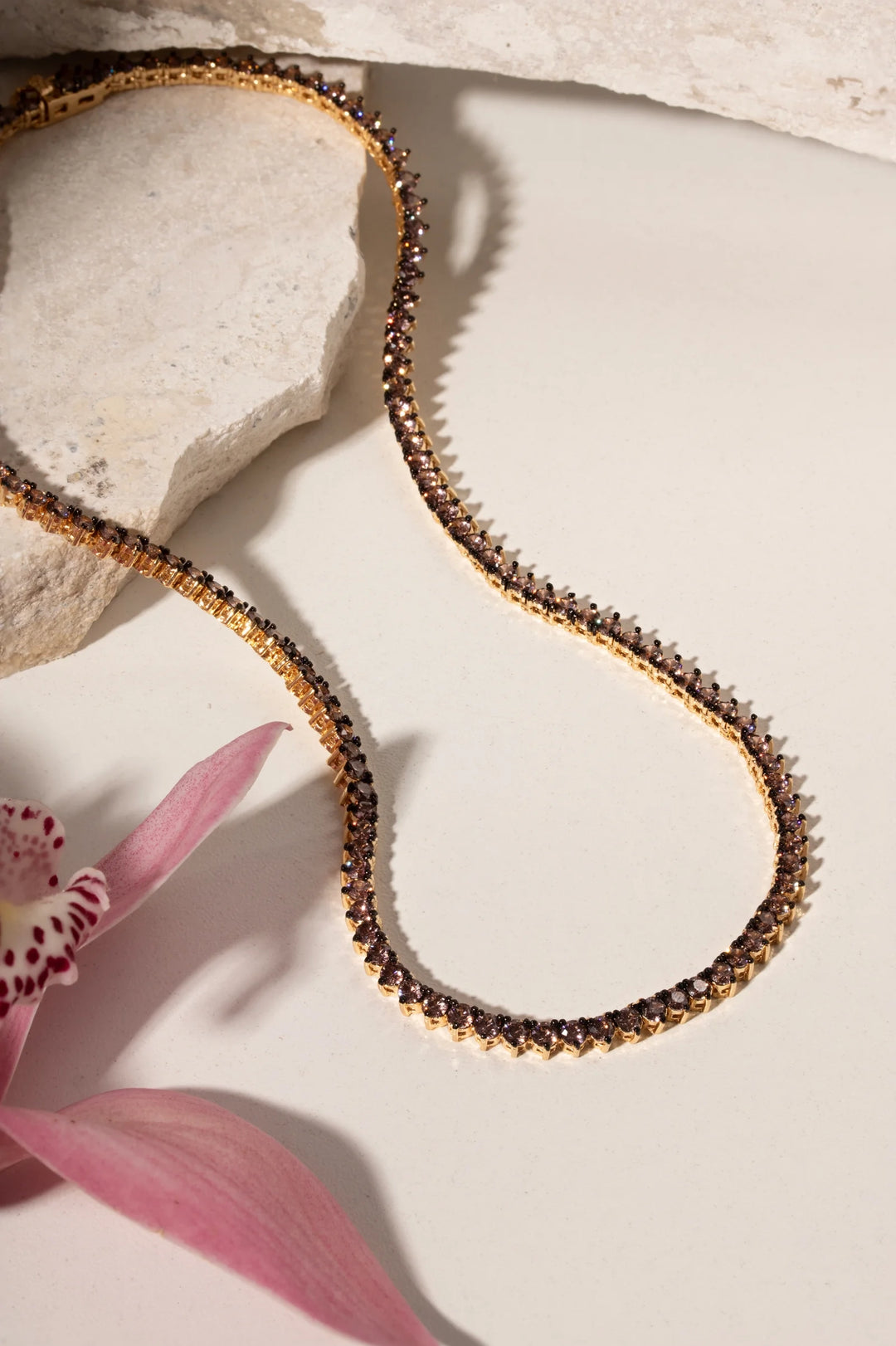 A delicate gold chain necklace with small dark brown or black beads, styled on a light cream surface with a pink orchid flower and textured stone props.