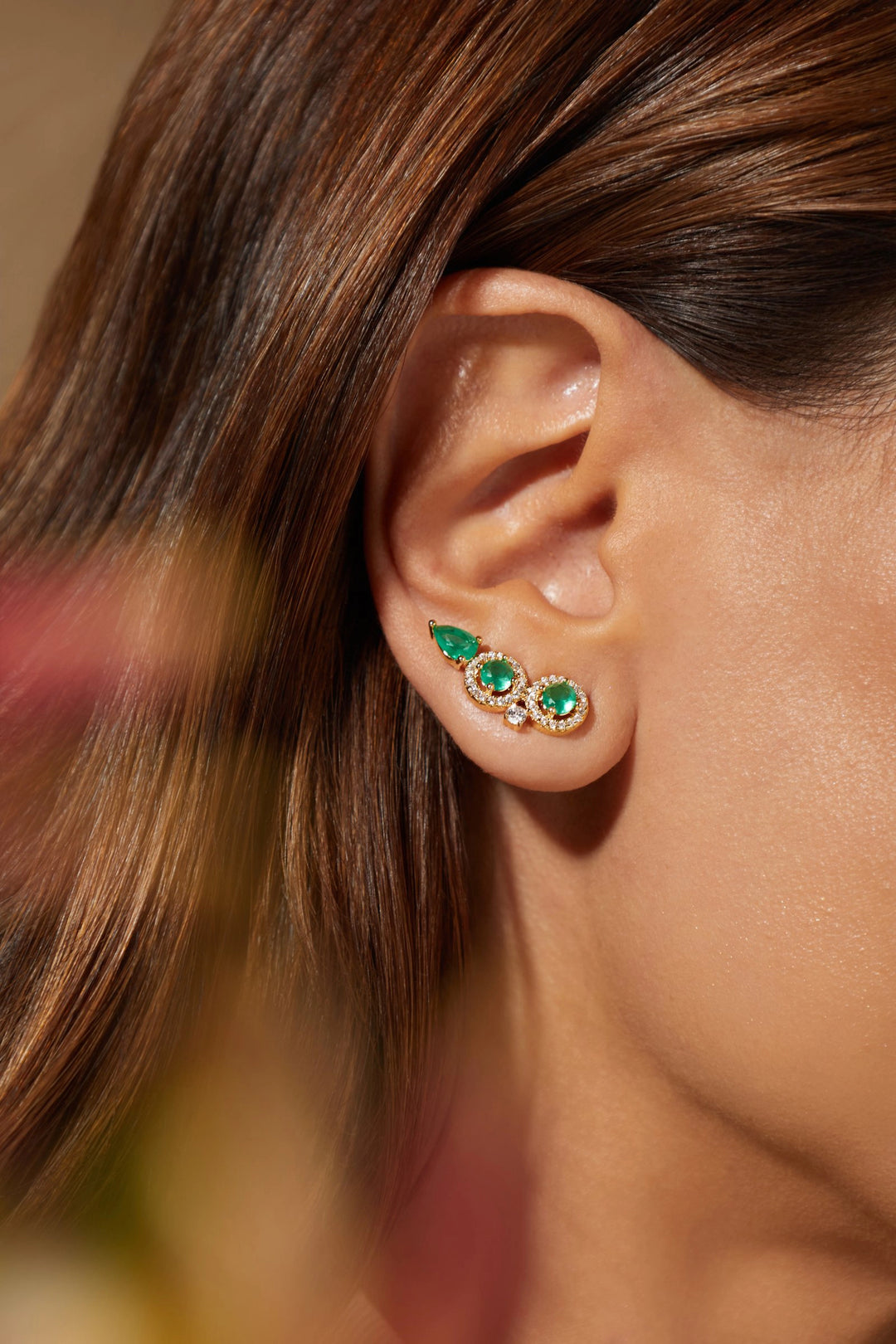 A close-up of a woman's ear wearing a pair of gold ear cuffs decorated with green emerald-cut gemstones and small clear crystals. 18k Gold Filled Elladora Australia