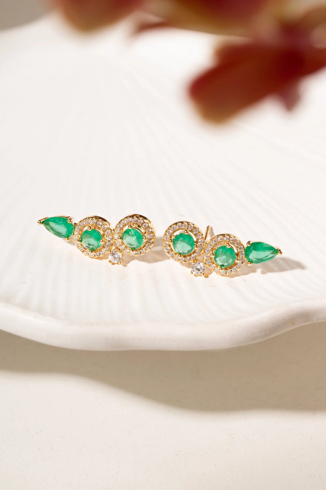A pair of gold-toned ear cuffs decorated with green gemstones and small clear crystals, resting on a white textured dish. 18k Gold Filled Elladora Australia