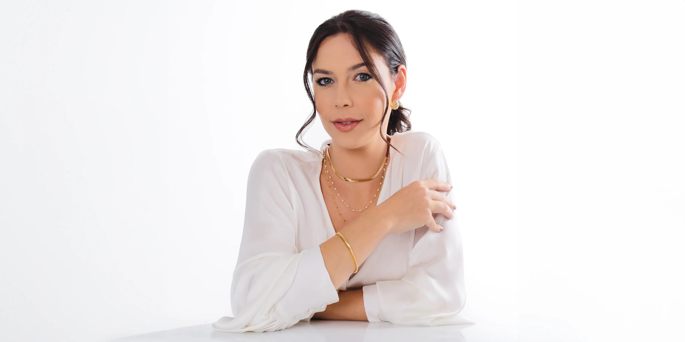 Elegant woman in a white blouse showcasing gold layered necklaces and hoop earrings from the latest jewellery collection.