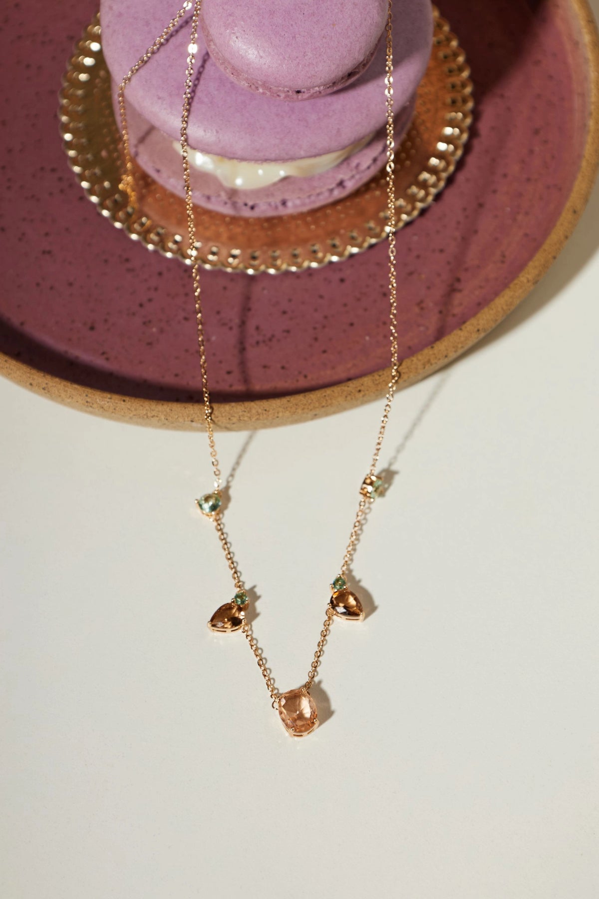 A delicate rose gold necklace with small heart-shaped charms and tiny green gemstone accents, draped over a purple macaron on a speckled purple plate against a white background.