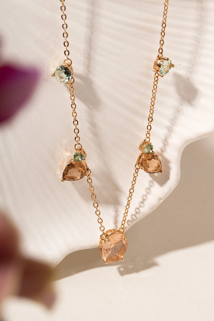 A delicate rose gold necklace with a Y-shaped drop design, featuring a large peach-colored teardrop gemstone at the center and smaller matching stones with light green accents on the sides, displayed on a white textured background.