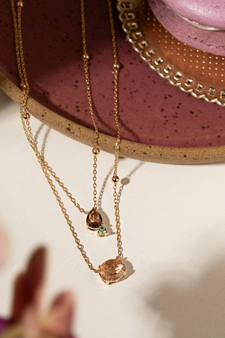 Two delicate rose gold chain necklaces with teardrop-shaped peach gemstones, displayed on a speckled mauve ceramic dish.
