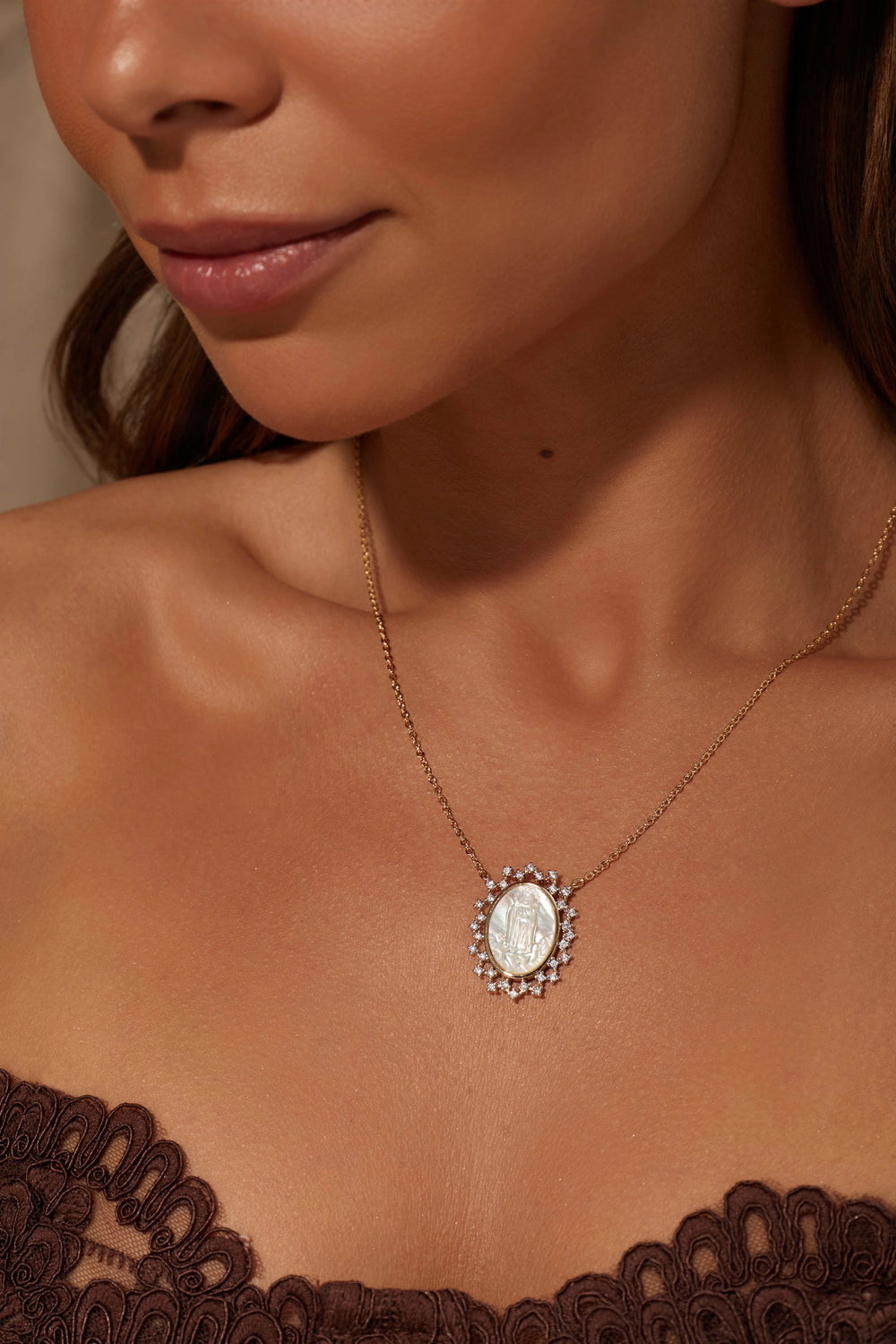 A close-up lifestyle shot of a model wearing a delicate gold chain necklace with an oval white pearl pendant surrounded by small clear crystals, styled with a brown lace top.