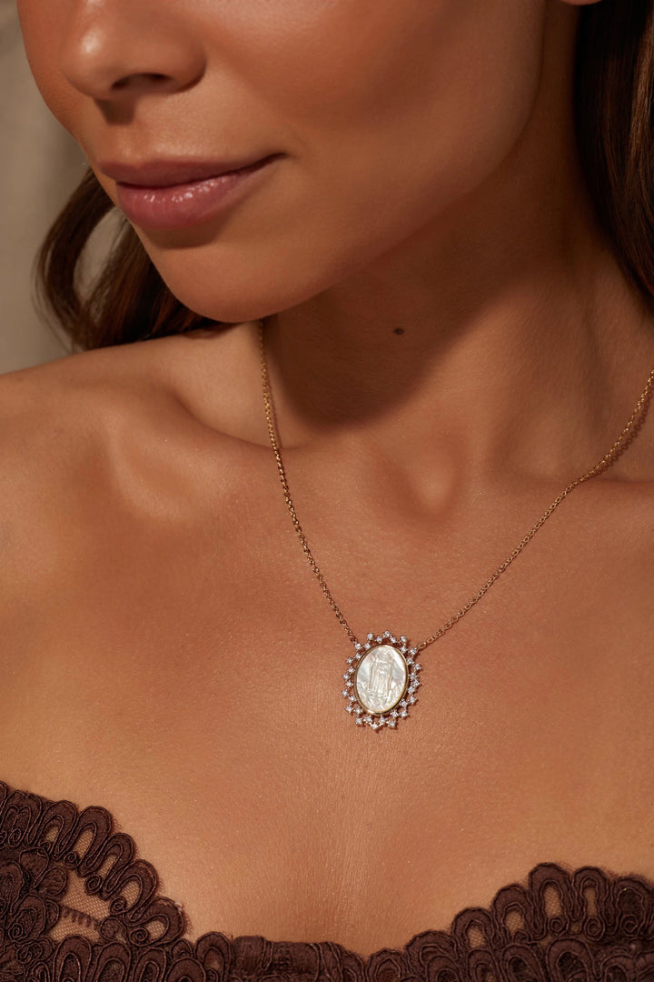 A close-up lifestyle shot of a model wearing a delicate gold chain necklace with an oval white pearl pendant surrounded by small clear crystals, styled with a brown lace top.