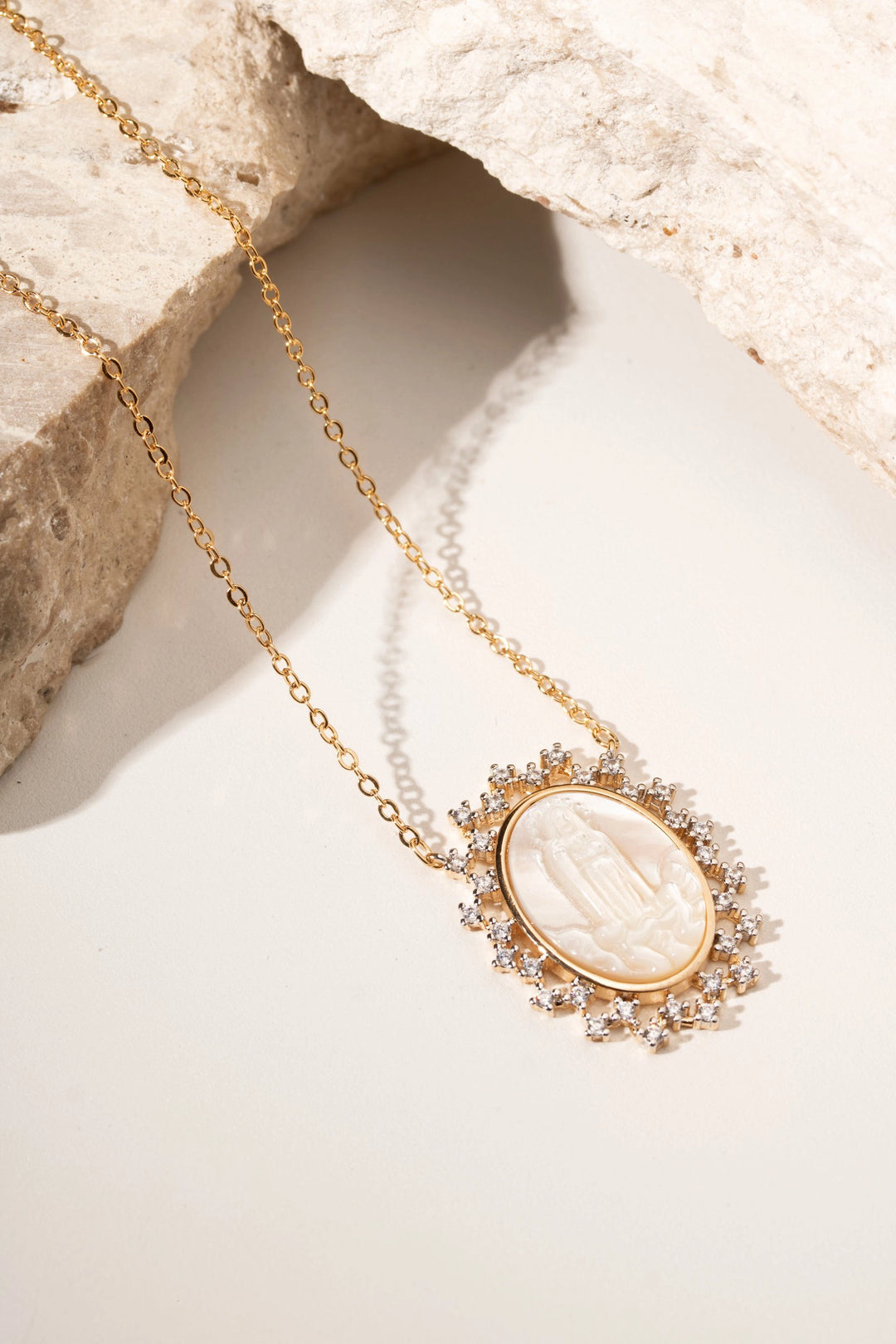 A gold pendant necklace featuring an oval carved white stone medallion surrounded by small clear stones, displayed on a light surface with a textured stone prop.