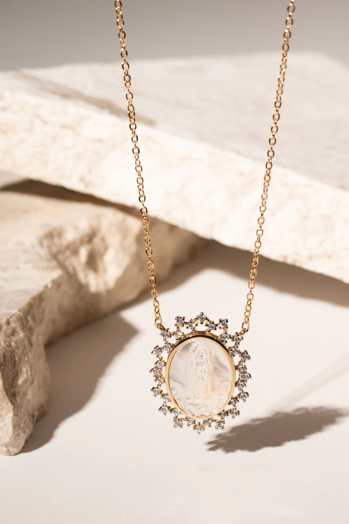 A gold chain necklace with an oval pendant featuring a white mother-of-pearl religious medallion surrounded by small clear stones, displayed on a light beige stone surface.