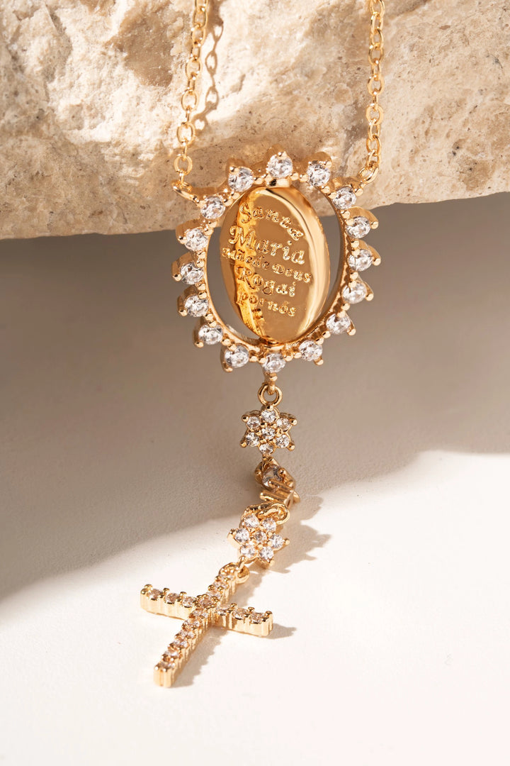A gold pendant necklace with a sunburst halo of clear crystals and a dangling cross charm, also accented with clear crystals. The oval center pendant features an engraved religious inscription.