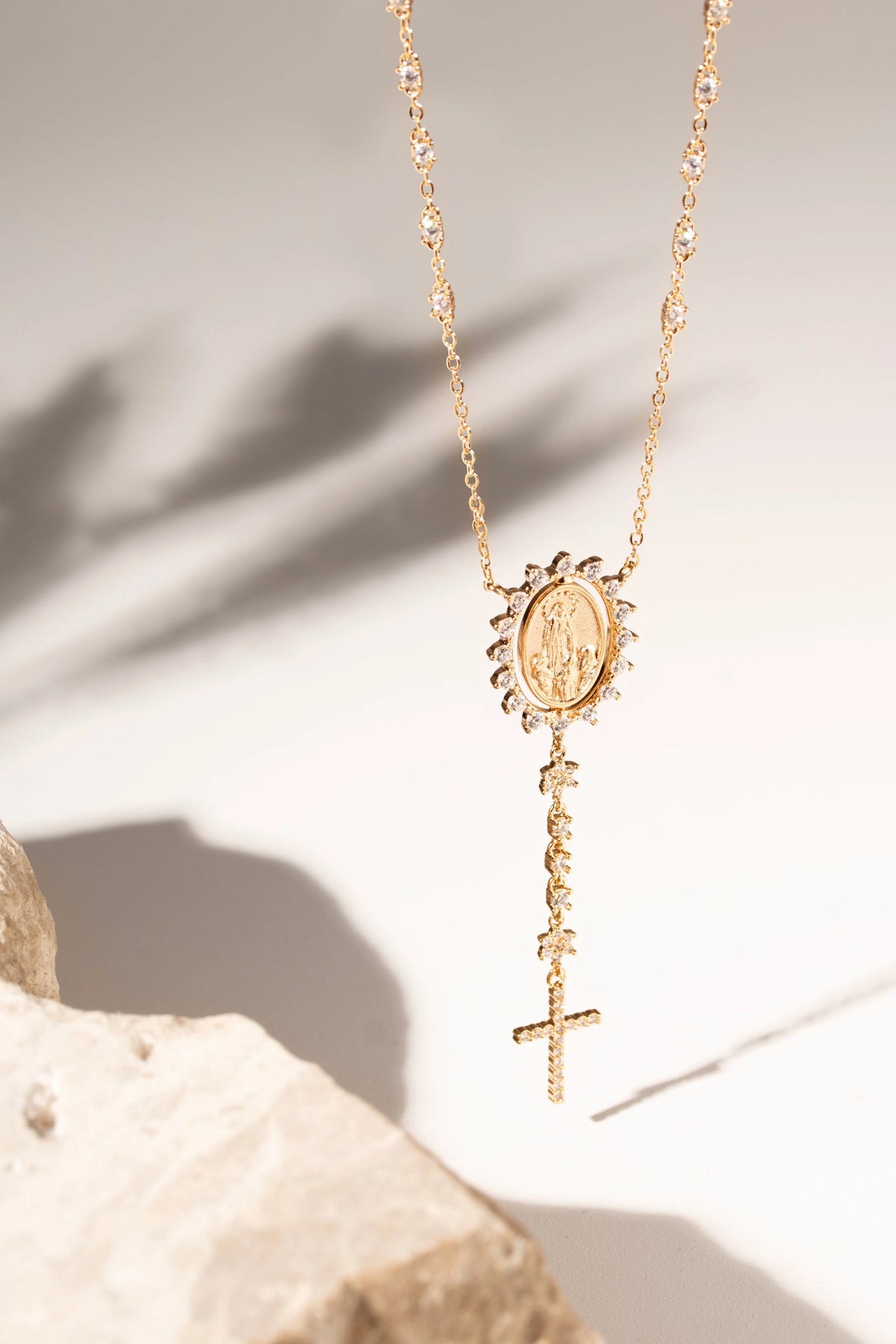 A gold-toned lariat-style necklace featuring a round medallion pendant and a dangling cross, displayed on a light neutral background with a stone prop and soft shadows.
