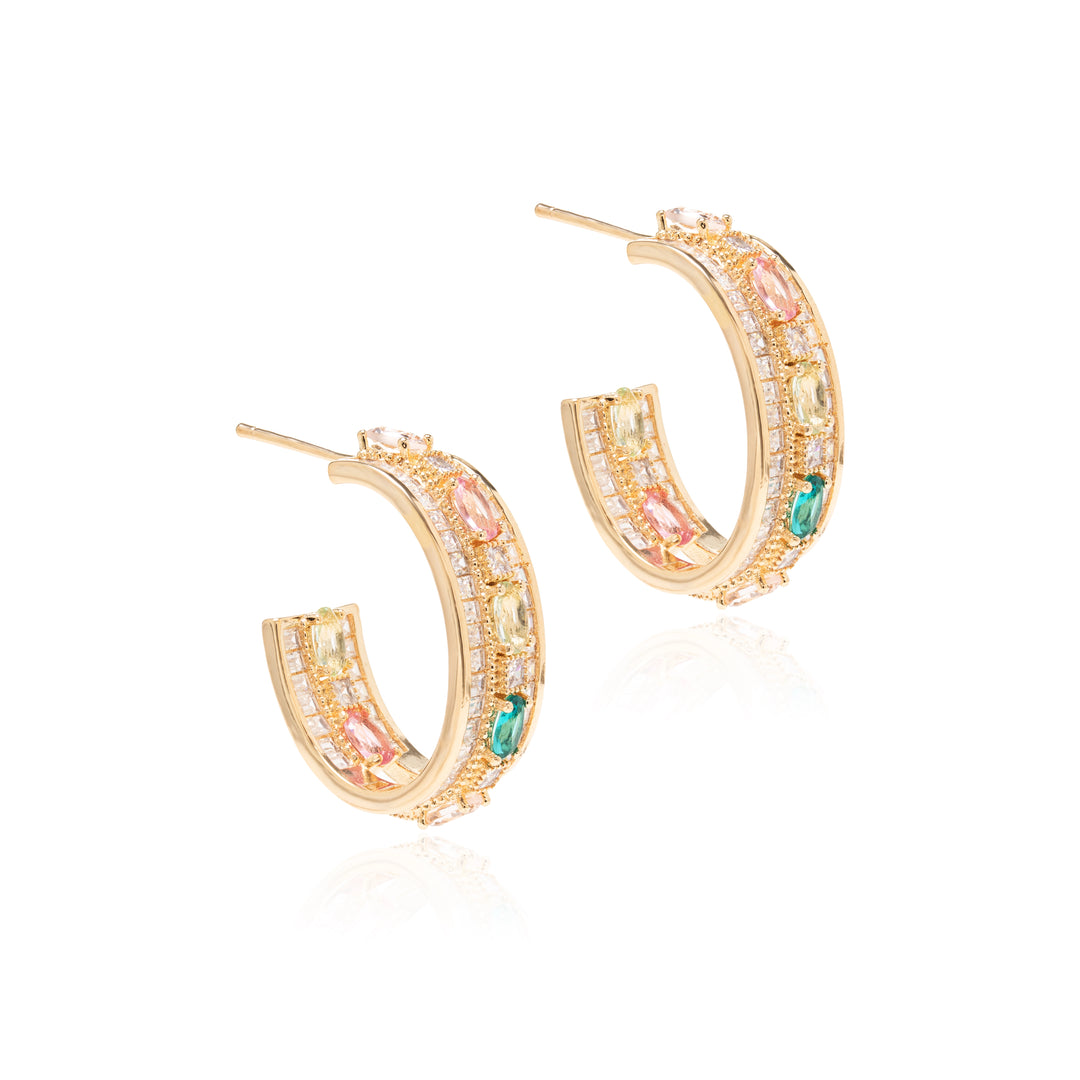 A pair of gold crescent hoop earrings decorated with pink, yellow, and green gemstones and pavé-set clear stones, shown on a clean white background with a soft reflection.