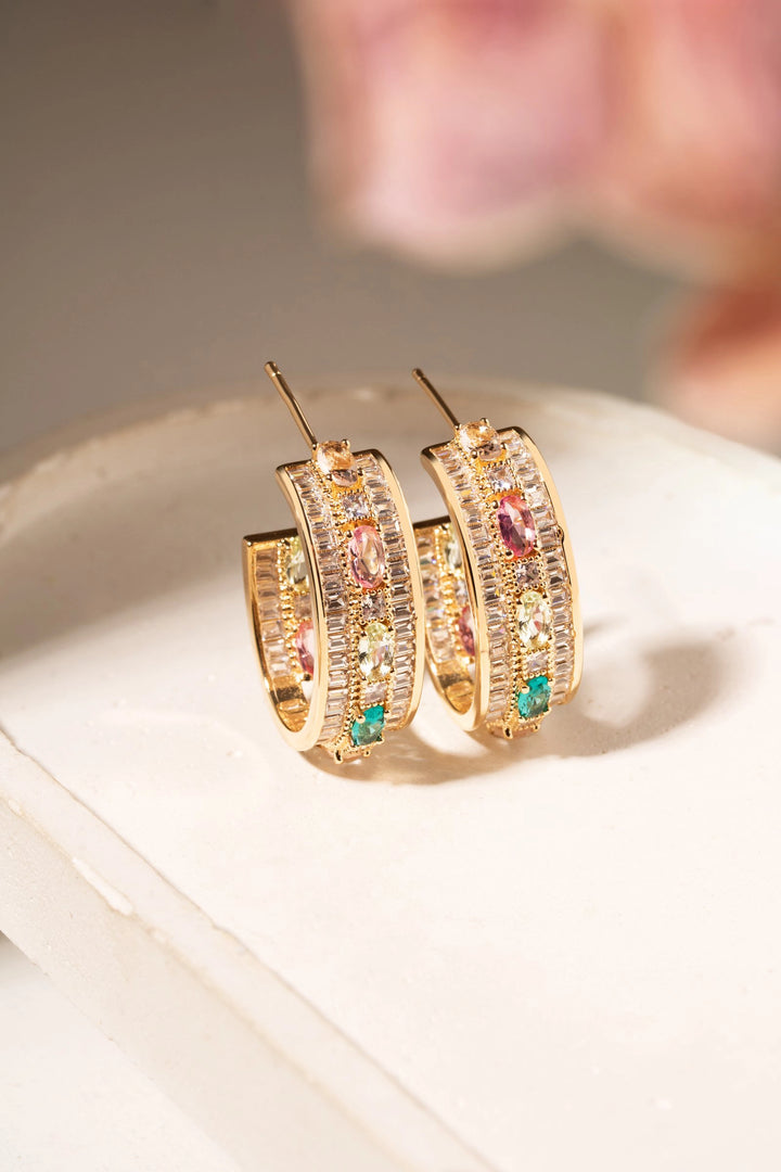 A pair of gold hoop earrings adorned with multiple rows of clear pavé-set stones and small colorful gem accents in pink, green, and orange, displayed on a white surface with a soft-focus pink background.