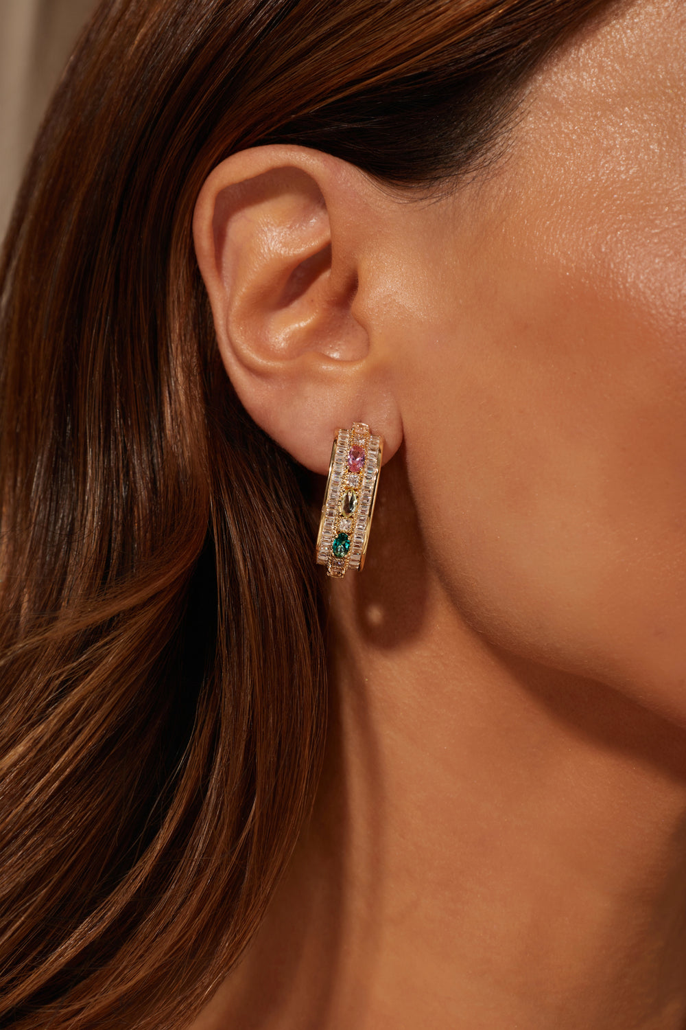 A close-up of a model's ear wearing a rectangular gold hoop earring adorned with small clear stones and colorful gem accents in pink, red, green, and blue.