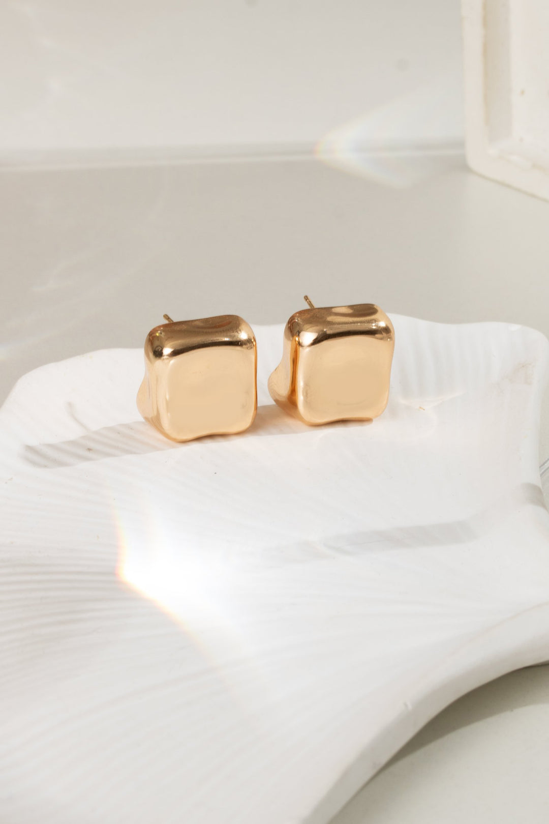 A pair of gold-toned cube stud earrings resting on a white textured ceramic dish against a bright, softly lit background.