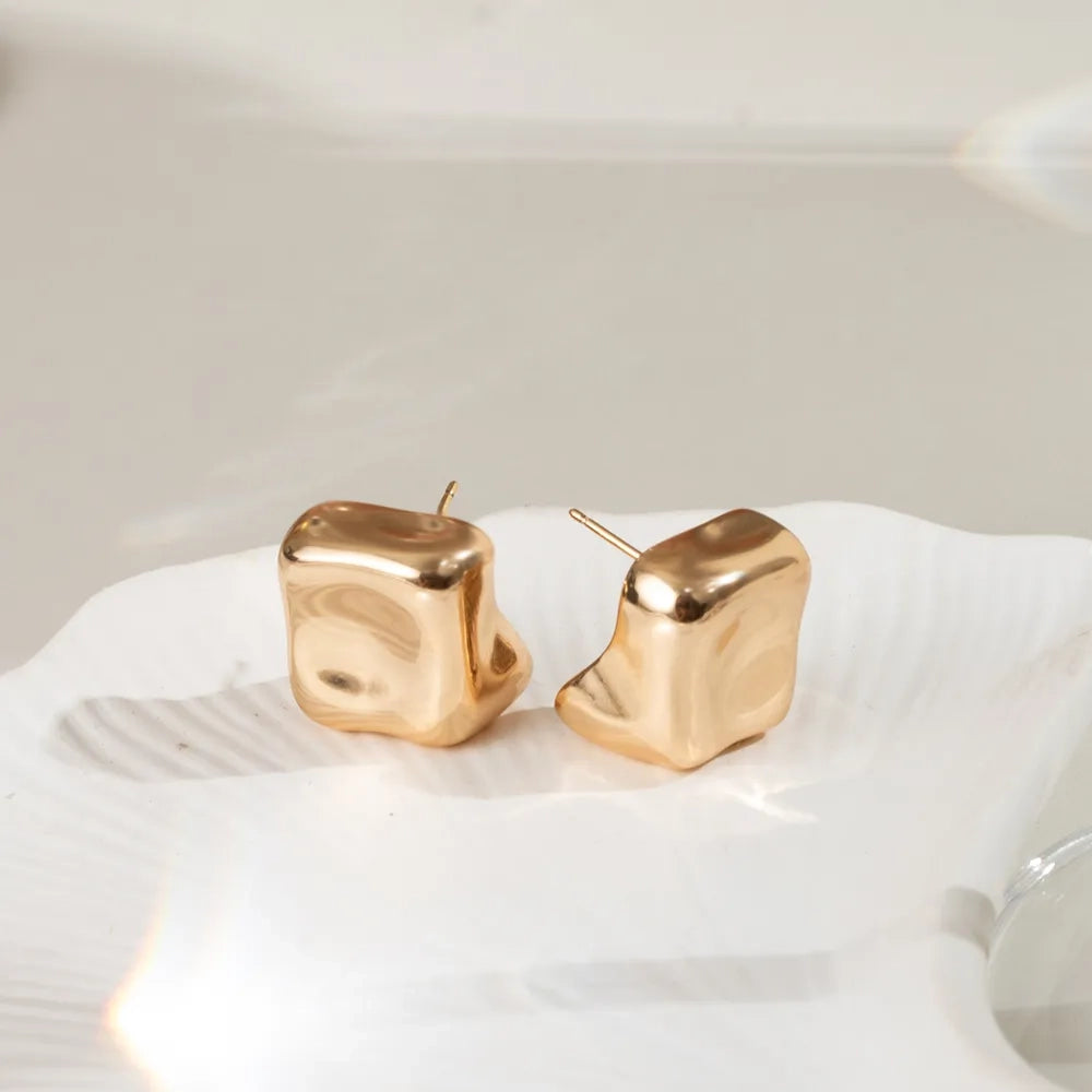 A pair of gold-toned, abstractly shaped stud earrings displayed on a white textured dish against a soft, light beige background.