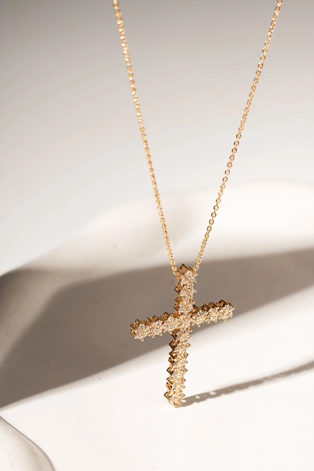 A gold cross pendant necklace with a textured, sparkling finish, displayed on a white surface with soft shadows.