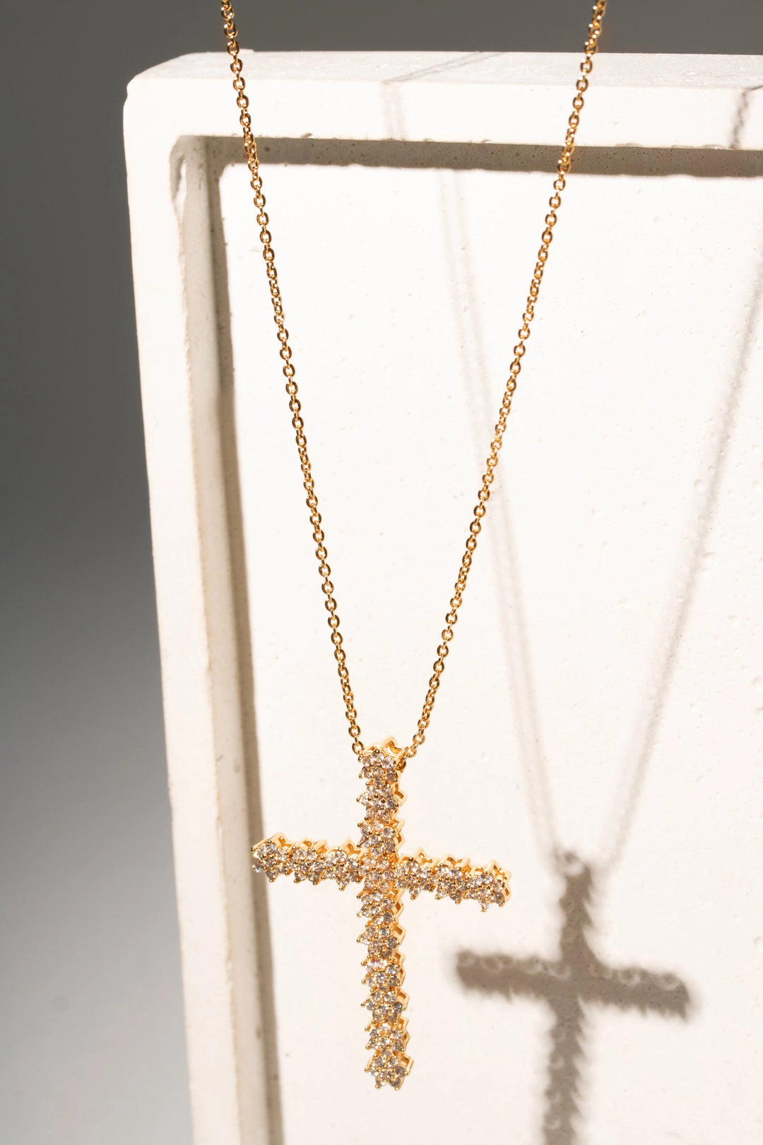 A gold cross pendant necklace encrusted with small clear stones, hanging from a delicate gold chain against a white display block.