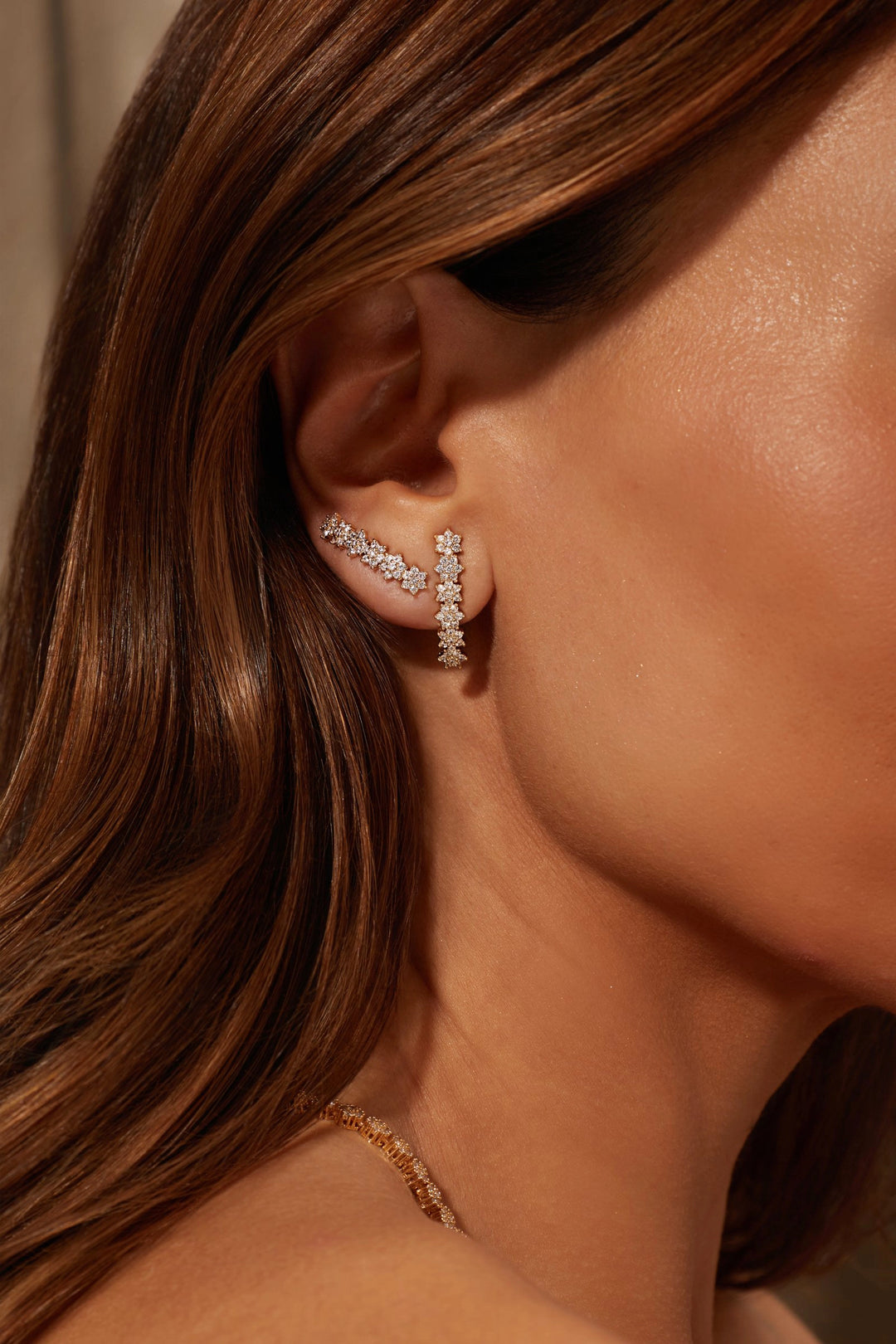 A close-up of a woman's ear wearing a delicate silver-toned ear cuff adorned with small clear stones. The warm, golden-toned lighting highlights her skin and hair, and a gold chain necklace is partially visible at the bottom.