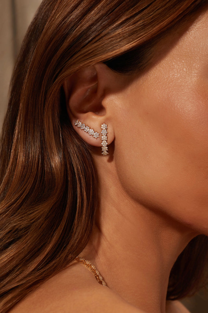 A close-up of a woman's ear wearing a delicate silver-toned ear cuff adorned with small clear stones. The warm, golden-toned lighting highlights her skin and hair, and a gold chain necklace is partially visible at the bottom.
