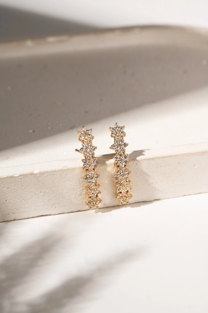 A pair of gold huggie hoop earrings adorned with clear pavé gemstones, resting on a textured beige stone surface with soft shadows.
