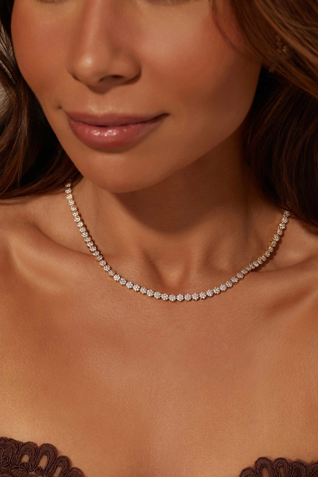 A close-up of a woman wearing a delicate diamond tennis necklace with a silver-toned setting, styled with a dark lace bralette.