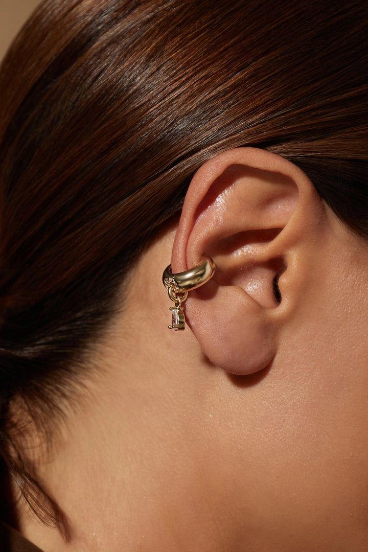 A close-up of a person's ear wearing a gold huggie hoop earring with a small dangling charm, set against dark brown hair and warm, direct lighting.
