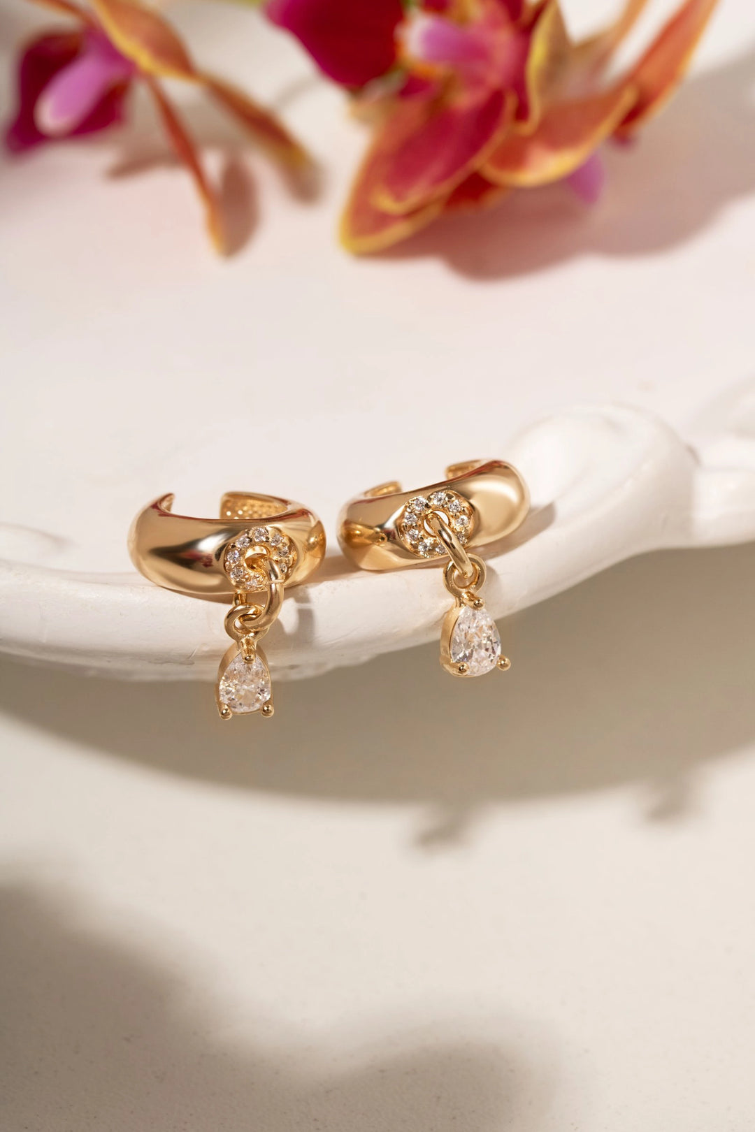 A pair of gold huggie hoop earrings with clear crystal accents and teardrop dangling charms, displayed on a white ceramic dish with blurred pink flowers in the background.