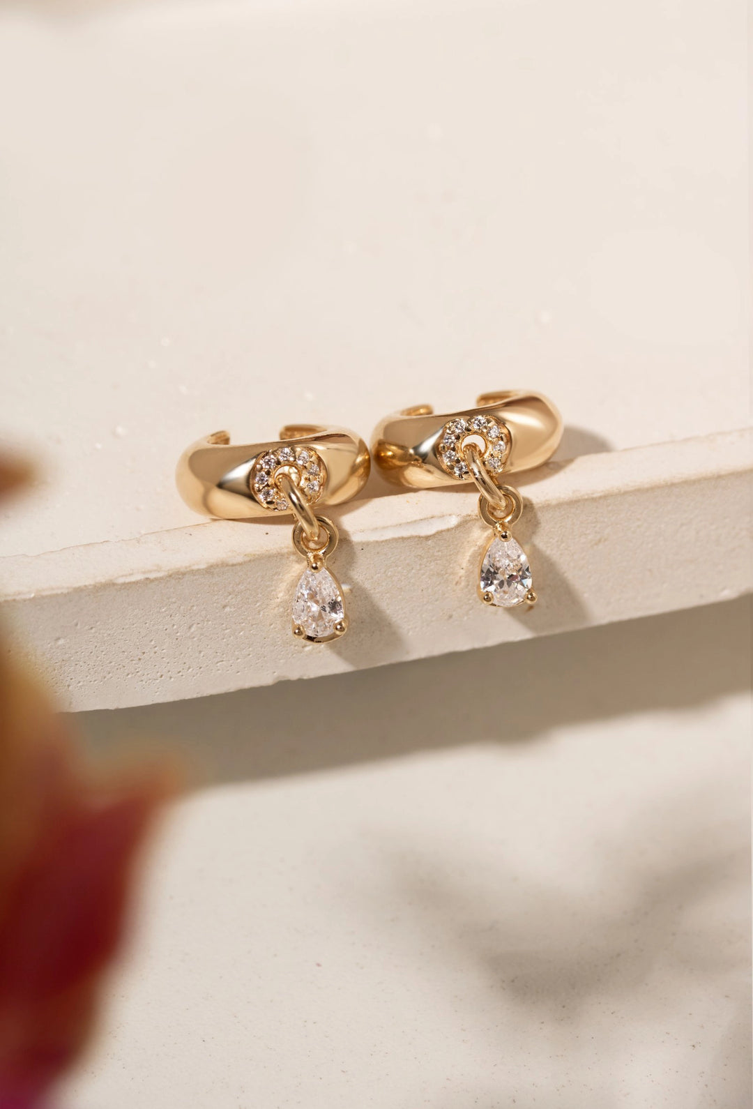 A pair of gold huggie hoop earrings with clear teardrop gemstones, displayed on a light beige textured surface with a soft-focus blurred background.
