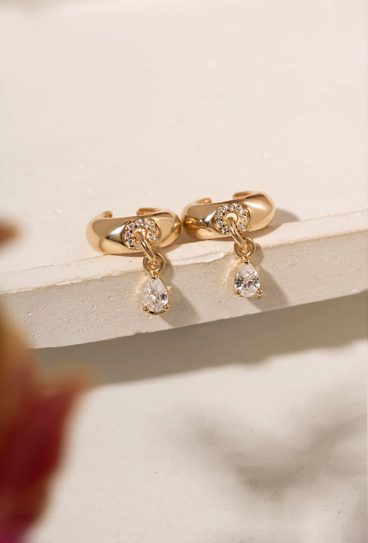 A pair of gold huggie hoop earrings with clear teardrop gemstones, displayed on a light beige textured surface with a soft-focus blurred background.