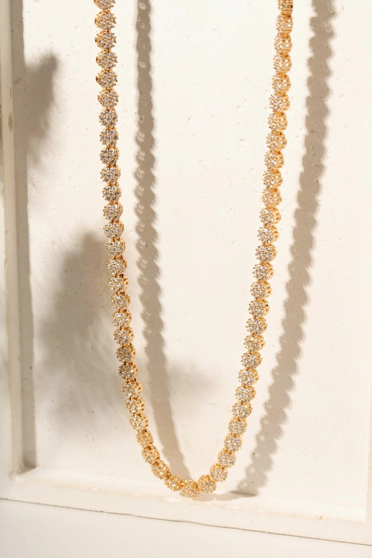A gold tennis-style necklace with clear rhinestones, draped in a U-shape against a white textured background with soft shadows.