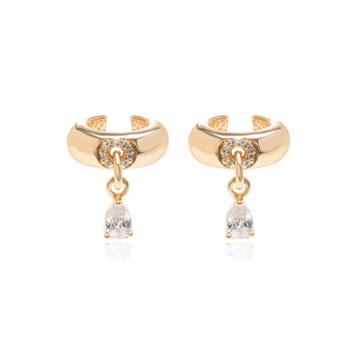 A pair of gold huggie hoop earrings with a dangling pear-cut clear gemstone, set against a solid white background.