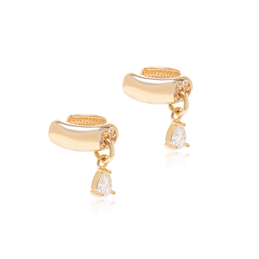 A pair of gold huggie hoop earrings with small clear stones along the front and a dangling pear-cut clear gemstone, shown on a white background with a soft reflection.