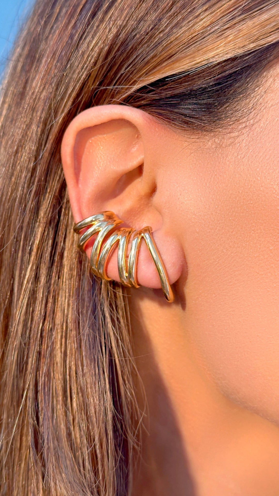 A close-up of a model's ear wearing a gold-toned, multi-band ear cuff with a twisted, spiral design.